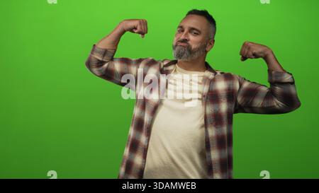 L'uomo ispanico di mezza età con capelli e barba grigi flette le braccia e grida in uno studio verde; sicurezza forza energia. Foto Stock