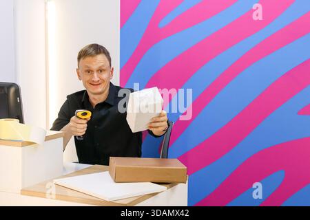 Uomo felice in camicia nera afferra lo scanner e pacco bianco al bancone con scatole, documenti, pareti vivaci. Foto Stock