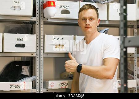 Il personale maschile mostra un gesto di approvazione rivolto verso la telecamera tra gli scaffali del magazzino Foto Stock
