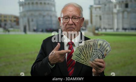 Un uomo anziano che ci tiene in mano dei dollari si trova di fronte alla torre di pisa e mostra ricchezza all'aperto. Foto Stock