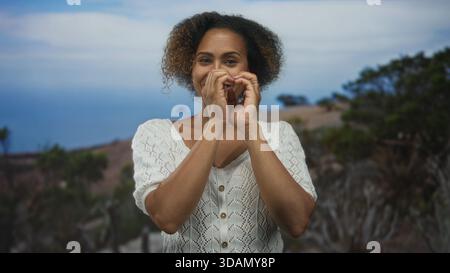 Donna che sorride e forma il cuore con le mani su una scogliera rocciosa che si affaccia sull'oceano; legame amoroso. Foto Stock