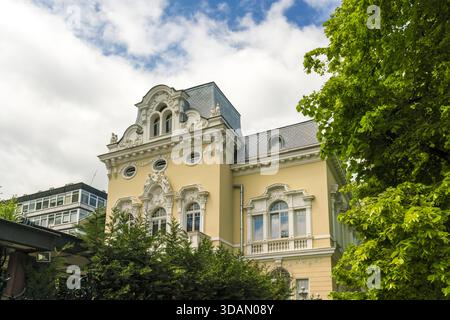 Vista di un edificio giallo ornato con finiture bianche, dettagli architettonici e alberi verdi su un cielo blu con nuvole, Sofia e Sofia Foto Stock