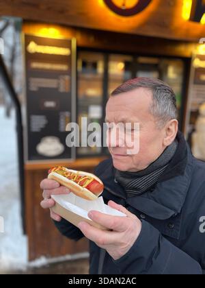 Uomo adulto che tiene l'hot dog vicino al chiosco di cibo di strada in inverno. Ambiente urbano all'aperto con luce calda. Foto Stock