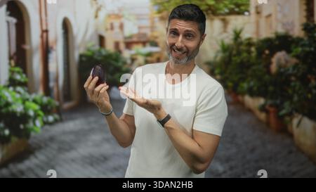 Uomo con avocado in mano e gesti verso di esso in una strada acciottolata; sano benessere allegro. Foto Stock