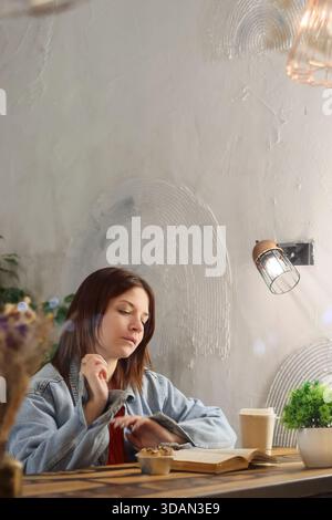 Giovane donna in giacca di denim che tocca il collo con attenzione mentre guarda un libro aperto in un accogliente ambiente da caffè Foto Stock