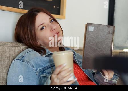 Giovane donna sdraiata con libro vicino al viso e tazza di caffè da asporto in un ambiente informale da caffe' Foto Stock