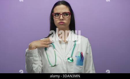 La dottoressa ispanica con uno stetoscopio si raffredda con le mani davanti a una parete viola, indossando occhiali e un'uniforme bianca, esprimendo calore Foto Stock