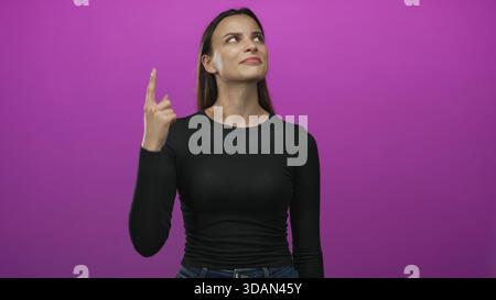 La donna punta il dito indice verso l'alto nello studio magenta mentre guarda in alto e indossa camicia e jeans neri; curiosità. Foto Stock