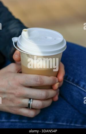 Persona in jeans che prende latte da asporto con coperchio bianco e schiuma visibile attraverso il contenitore trasparente su sfondo sfocato Foto Stock