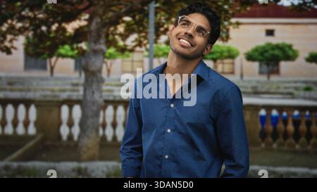 L'uomo che indossa occhiali e una camicia blu aperta sorride, inclina la testa e mostra il collo davanti al cortile dell'edificio con balaustra e alberi; tranquillità e tranquillità Foto Stock