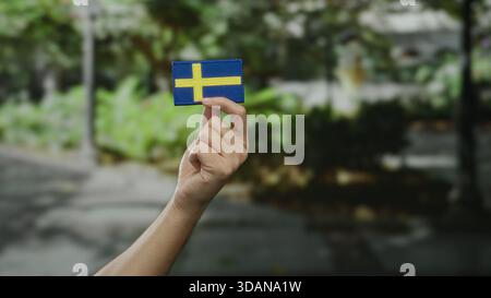 Mano che regge la bandiera svedese in un ambiente cittadino all'aperto che simboleggia l'orgoglio nazionale e la cultura. Foto Stock
