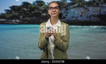 Donna con occhiali da vista, utilizza lo smartphone tenendo in mano il dispositivo e guardando in basso in uno studio con sfondo da spiaggia; concentrazione. Foto Stock