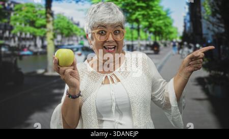 Donna che tiene la mela verde in mano sinistra e punta con la mano destra in strada, sorride con i capelli grigi e indossa gli occhiali; gioiosa curiosità. Foto Stock