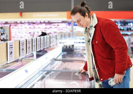 uomo in giacca rossa che sceglie il cibo surgelato al supermercato. Cliente maschio che guarda nel cassettiere del congelatore presso il supermercato. Foto di alta qualità Foto Stock