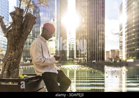 Giovane uomo nero in un abbigliamento casual, che si concentra sul suo smartphone mentre si appoggia a un albero, con grattacieli moderni e acqua scintillante nella luminosa e Foto Stock