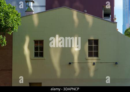 La luce del sole proietta ombre drammatiche e ondulate sulla facciata di un edificio verde pallido, caratterizzato da due finestre a griglia, tra varie strutture urbane. Foto Stock