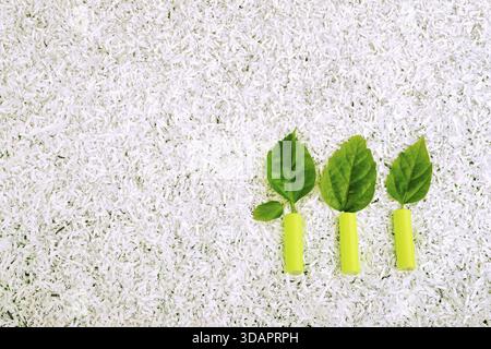 Tre piccole piante fatte di batterie verdi e foglie fresche su un mucchio di carta bianca triturata Foto Stock