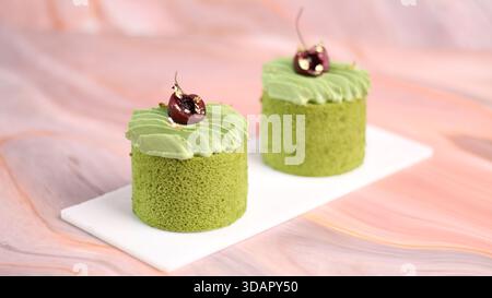 Due mini torte di tè verde matcha piacevoli, elegantemente condite con panna vorticosa, ciliegia tagliata a metà e foglia d'oro commestibile Foto Stock