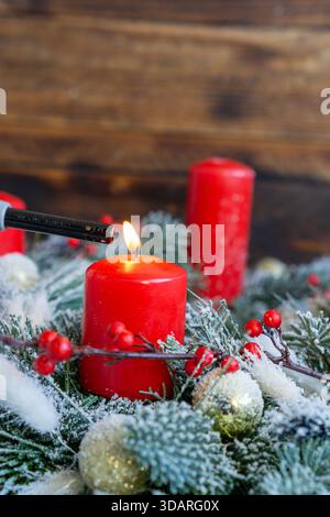 Una scena ravvicinata mostra una candela a colonna rossa accesa tra rami sempreverdi smerigliati, bacche rosse brillanti e ornamenti dorati. Foto Stock