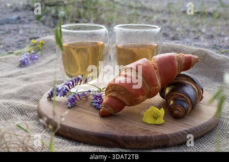 colazione con croissant, tè caldo in tazze di vetro e fiori su una tavola di legno su tessuto burlap testurizzato Foto Stock