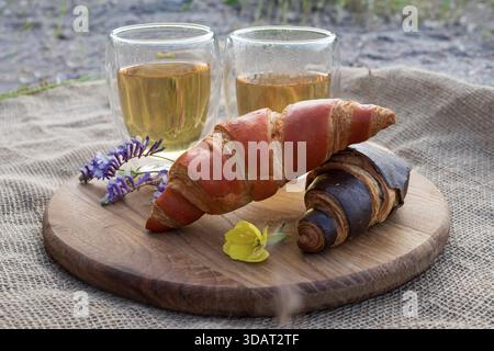 colazione con croissant, tè caldo in tazze di vetro e fiori su una tavola di legno su tessuto burlap testurizzato Foto Stock