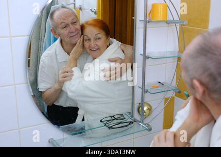 Coppia senior felice che fa routine di bellezza quotidiana in bagno a casa, sposi maturi sorridenti che abbracciano e guardano allo specchio, si preparano insieme la mattina, godendo della cura del corpo. Foto Stock