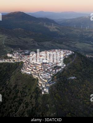 Vista aerea di un villaggio dalle sfumature dorate aggrappato a una ripida collina, coronato da una chiesa pallida, sullo sfondo di montagne scure e turbine eoliche, R Foto Stock
