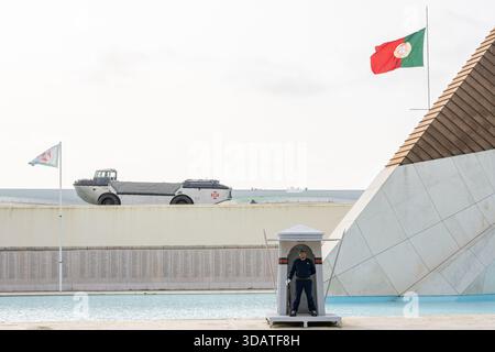 Lisbona Portogallo 4 dicembre 2025. Una guardia militare portoghese si trova a un posto mentre un veicolo militare anfibio è esposto dietro i Combatentes Foto Stock