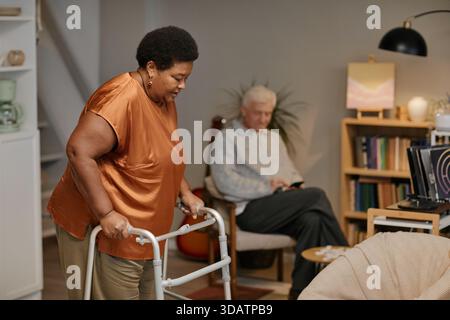 Donna nera anziana che usa Walker mentre l'uomo caucasico anziano seduto sullo sfondo Foto Stock