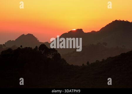 View of dark, shadowy mountains silhouetted against a vibrant, fiery sunset, where the sun dips below the peaks, casting an orange glow, Islamabad, Is Foto Stock