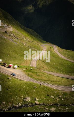Vista di una strada tortuosa che serpeggia attraverso lussureggianti colline verdi, auto che si muovono lungo il sentiero, illuminate dai raggi del sole, Poienari, Transilvania, Romania. Foto Stock