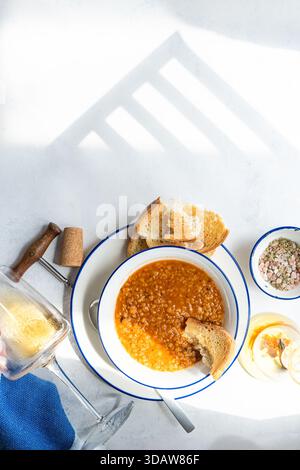 Una calda ciotola di zuppa di lenticchie greca servita con pane croccante e un bicchiere di vino bianco. L'ambiente illuminato dal sole mette in risalto i vibranti colori della tradizione Foto Stock