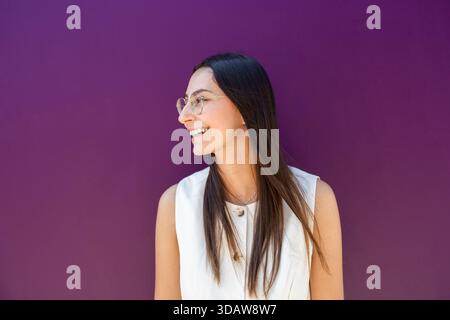 Donna d'affari in abito bianco si erge con sicurezza contro una vivace parete viola, sorridendo mentre guarda di lato. Lo sfondo colorato migliora la sua p Foto Stock