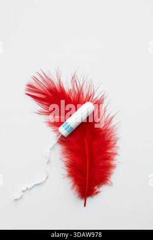 Un tampone bianco si trova accanto a una piuma rossa vivace su uno sfondo bianco, che simboleggia la salute mestruale, la femminilità e i cicli naturali con un'ar sottile Foto Stock
