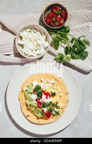 Vista dall'alto di Huevos Rancheros strapazzato su una tortilla con guacamole, formaggio e pomodori su un piatto beige. Contorni di salsa e formaggio su un panno bianco Foto Stock
