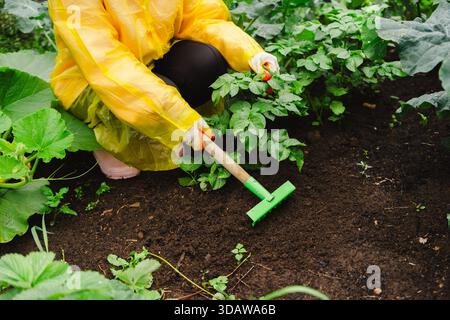 Una donna con un impermeabile giallo tende alle sue piante in un giardino lussureggiante. Circondata dal verde, usa un piccolo rastrello per coltivare il terreno, promuovendo Foto Stock