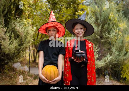 Due allegre ragazze vestite da streghe in costumi vivaci celebrano Halloween all'aperto. Uno contiene una zucca, mentre l'altro porta un calderone, embod Foto Stock