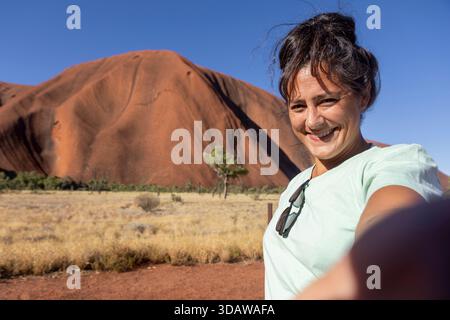 Una allegra donna turistica sorride molto, scattando un selfie mentre visita l'iconica Uluru in Australia. Il monolite di arenaria rossa si erge maestosamente a Foto Stock