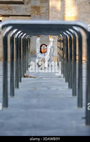 Una donna asiatica con un abito in argento riflettente posa artisticamente sotto una serie di barre metalliche in un ambiente urbano, trasudando fiducia e stile moderno Foto Stock