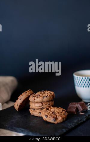Su una superficie di ardesia scura viene esposta una pila di biscotti fatti in casa carichi di gocce di cioccolato. Accanto ai biscotti ci sono pezzi di cioccolato, con un motivo Foto Stock