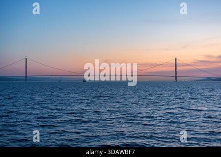 Il ponte sospeso di Canakkale attraversa elegantemente l'acqua, sagomato contro un cielo vibrante al tramonto, che riflette le sfumature di arancione e rosa, creando un ambiente sereno Foto Stock