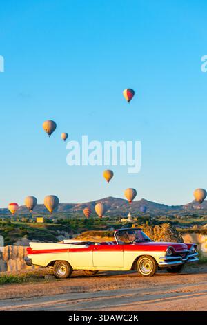 Una classica auto a noleggio decappottabile parcheggiata in Cappadocia all'alba con vivaci palloni ad aria calda che adornano il cielo, creando una scena pittoresca per il tour Foto Stock
