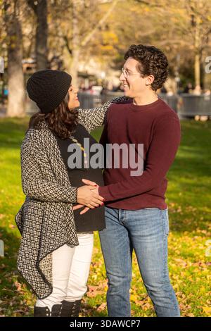 Una donna incinta gioiosa e la sua compagna abbracciano, condividendo un momento intimo circondato da foglie autunnali in un parco soleggiato, celebrando l'amore e il nuovo begi Foto Stock