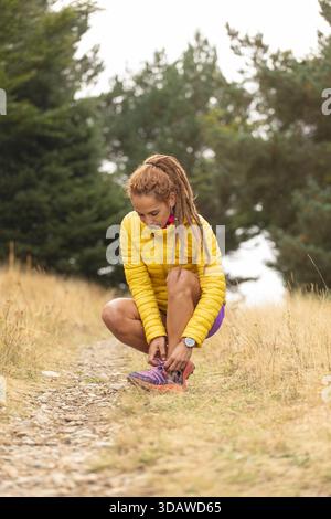 Una donna con una giacca gialla accovacciata su un sentiero roccioso, legando le sue scarpe. Si sta preparando per una pista panoramica con lussureggianti alberi verdi che la circondano Foto Stock