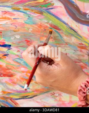 Una donna dipinge sapientemente una vibrante scena astratta su una grande tela, unendo colori ricchi per creare un'opera d'arte dinamica e suggestiva. La sua concentrata espr Foto Stock