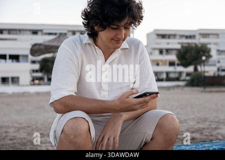 Un giovane uomo si siede su una spiaggia al tramonto, a controllare il suo cellulare. Indossando un abbigliamento casual, è rilassato e si gode il tranquillo ambiente all'aperto vicino Foto Stock