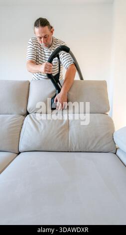 Un uomo maturo pulisce il suo divano di casa utilizzando una macchina che utilizza acqua preionizzata. Questo metodo ecologico evita le sostanze chimiche e assicura al contempo il lettino Foto Stock