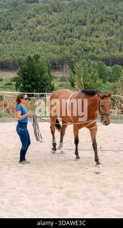 Una donna con una camicia blu conduce un cavallo marrone con cura su una pista sabbiosa, circondata da vegetazione lussureggiante e alberi, mostrando un sereno allenamento equestre Foto Stock