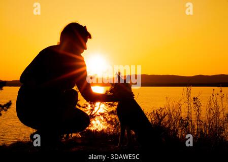 Una donna accovacciata accanto a un cane, caratterizzata da un brillante tramonto su un lago tranquillo. La scena cattura un momento sereno nella natura, perfetto per Foto Stock