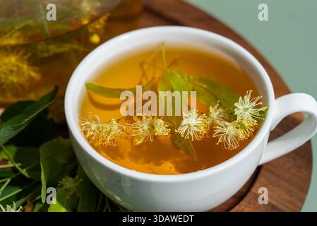 Primo piano di una rilassante tazza di tiglio alle erbe presentata in una tazza bianca, decorata con fiori freschi di tiglio, su sfondo verde. Il tè riposa Foto Stock